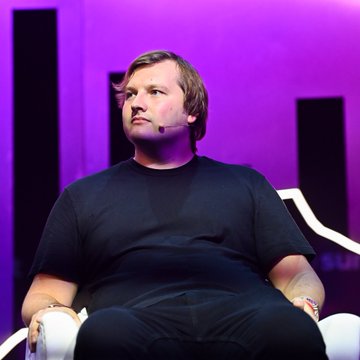 In the image, there's a person sitting down with their hands clasped together in front of them. The scene appears to be set up as if for a presentation or interview by someone named Stani Kulechov. He seems deep in thought while looking off into the distance. The atmosphere feels serious and focused as he contemplates something, possibly related to his work or personal life.