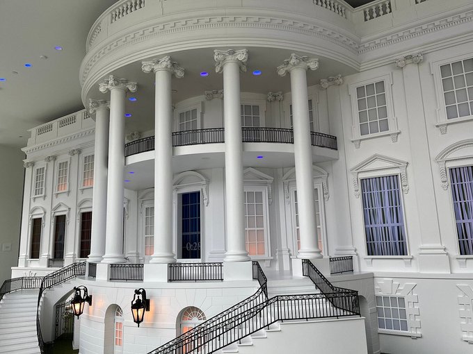 An ornate white interior resembling the White House, featuring grand Corinthian columns, curved double staircases, classical architectural details, and dramatic blue and orange accent lighting.