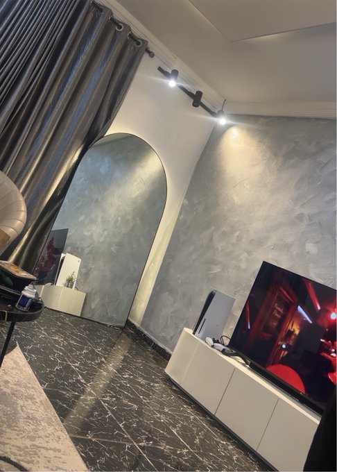 A modern interior room features textured grey walls, a large flat-screen TV on a white stand, marble-patterned flooring, and dark curtains.