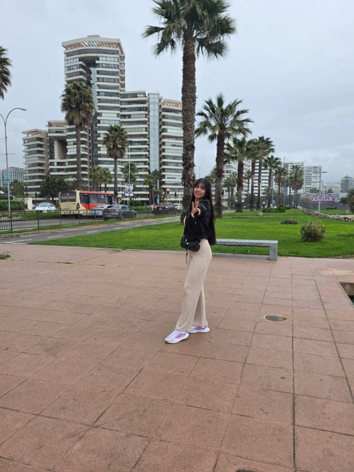 A young woman poses on a paved walkway near palm trees and modern city buildings, wearing a black top, beige pants, and white sneakers while carrying a black bag.