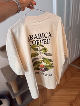 A cream-colored long-sleeve shirt hanging on a wooden hanger, featuring a botanical illustration of a coffee plant with the text 'ARABICA COFFEE' and 'ART OF FLORA'.