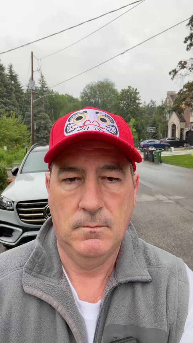 The image shows a man wearing a red baseball cap with an animal face design standing next to his car. He appears to be posing for a photo, possibly for a social media post or advertisement. The background of the image features a street scene with cars parked along the side and trees in the distance.