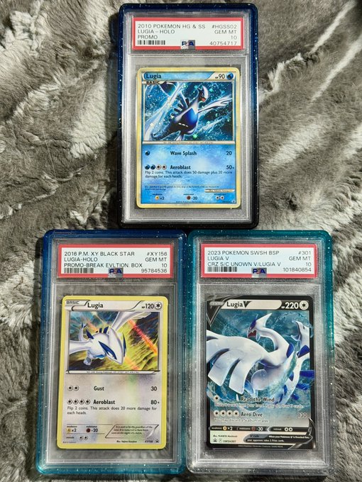 Three PSA-graded Lugia Pokémon cards are displayed on a gray fluffy surface: a 2010 HG&SS Lugia Holo (Gem Mint 10), a 2016 XY Black Star Promo Lugia Holo (Gem Mint 10), and a 2023 SWSH BSP Lugia V (Gem Mint 10).