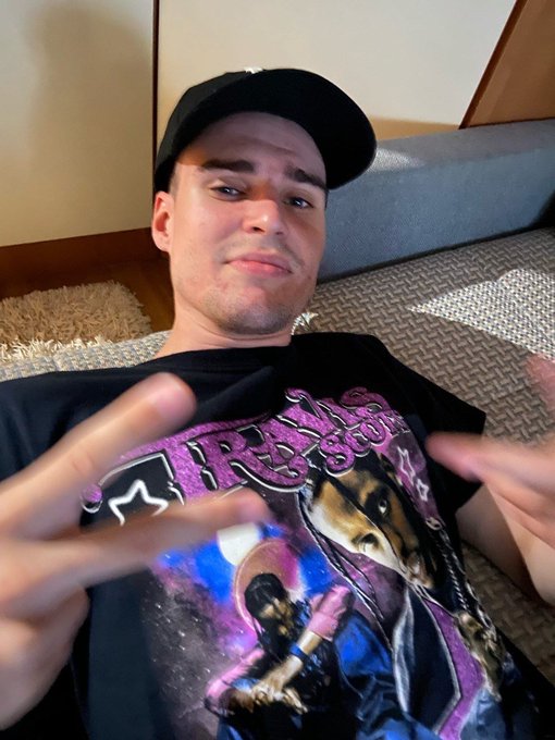 A selfie of a young man wearing a black cap and a black graphic t-shirt, making a peace sign with his fingers on a couch.