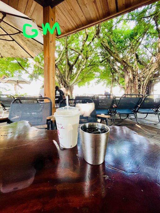 An outdoor cafe table holds a plastic cup and a stainless steel tumbler, with the letters 'GM' drawn in green in the upper left corner.