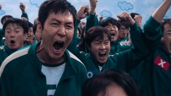 An intense scene from the TV show Squid Game showing contestants in green tracksuits cheering with raised fists and open mouths, including one with an "X" patch on their jacket.