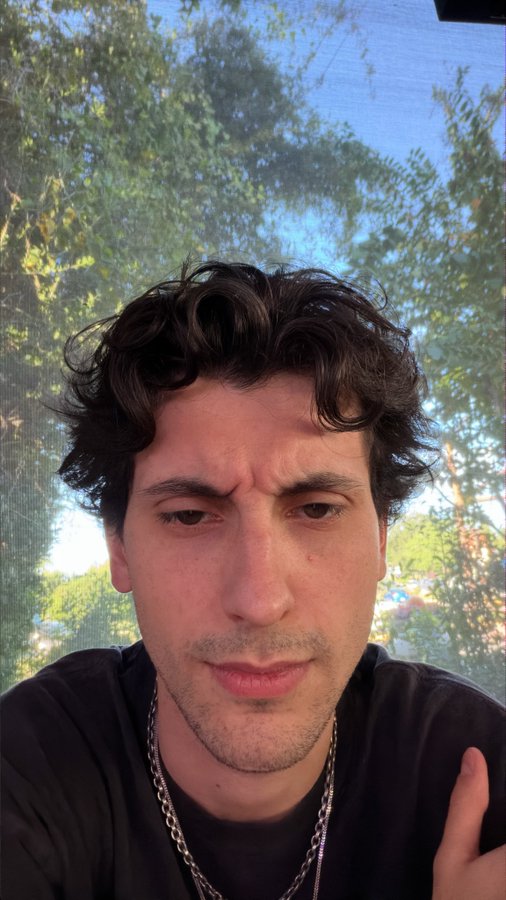 A young man with dark curly hair takes a close-up selfie outdoors, surrounded by green foliage. The image shows no visible text or numbers, and doesn't directly relate to the caption about a "dickbuttify" pump fun mobile app account.