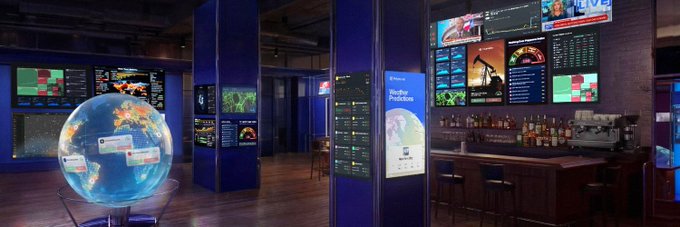 A modern digital trading lounge features multiple Bloomberg terminals displaying financial data, world maps, and market charts alongside a coffee bar and a glowing interactive globe displaying locations like "Mali." The image complements the post's theme by visually evoking a high-tech, data-driven trading environment, suggesting Polymarket's new perpetual futures (perps) offering—complete with up to 20x leverage—enters a sophisticated space typically associated with traditional professional trading.