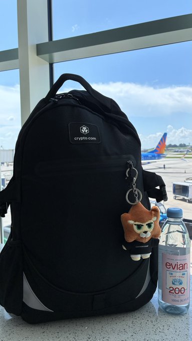 A black crypto.com backpack with a small sloth keychain sits at an airport gate next to a bottle of Evian water, with a Southwest Airlines plane visible through the window.