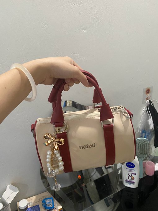 A hand holds up a small cream and burgundy handbag with a pearl and gold bow charm and the brand name 'natoli' on it.