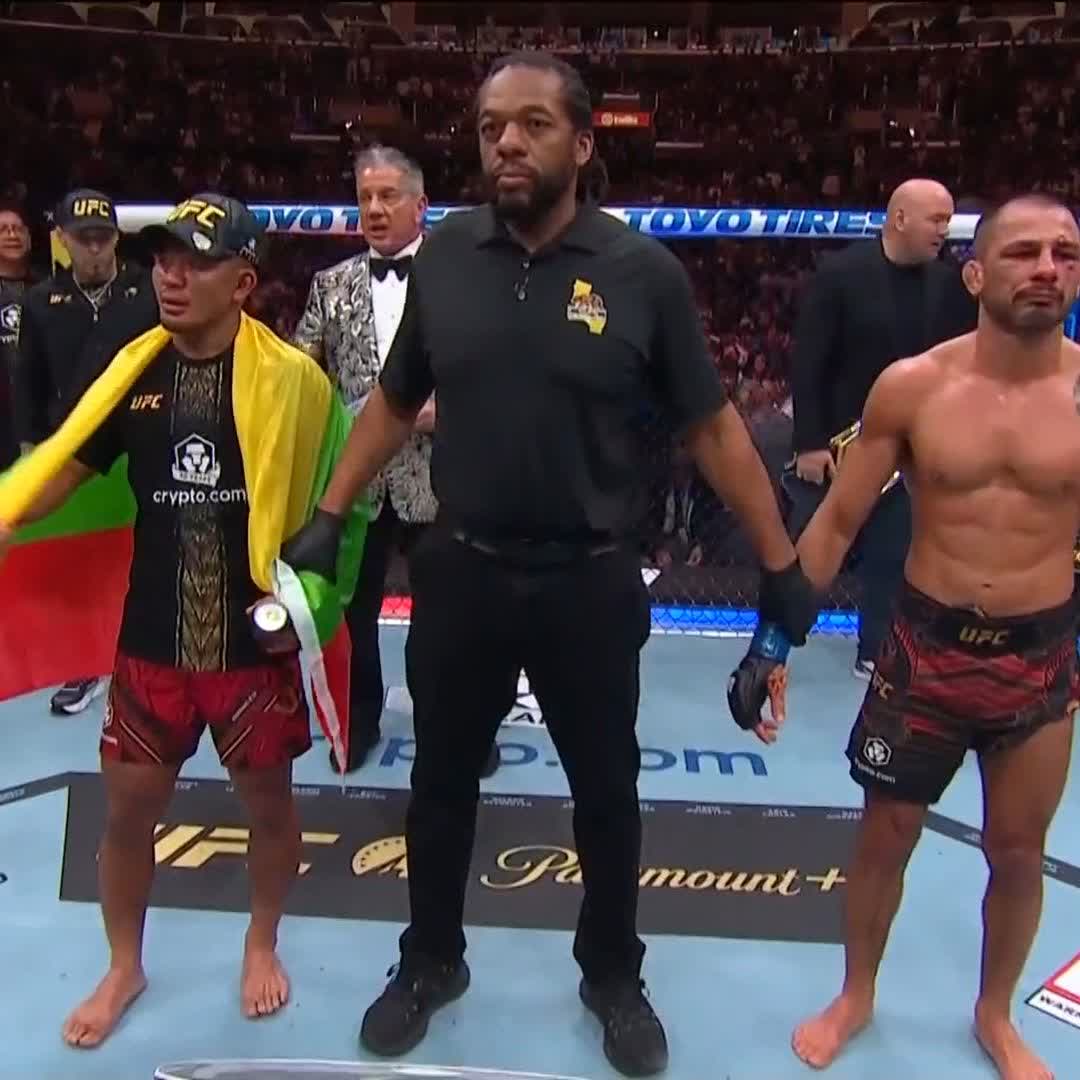 A referee raises the hands of two fighters in a UFC octagon, with the fighter on the left wearing a cap and draped in a Lithuanian flag next to the UFC and crypto.com-branded belt. The image captures the post-fight moment referenced in the caption, where Joshua Van is announced as the still champion after retaining his belt by unanimous decision at UFC 331, broadcast on Paramount+.