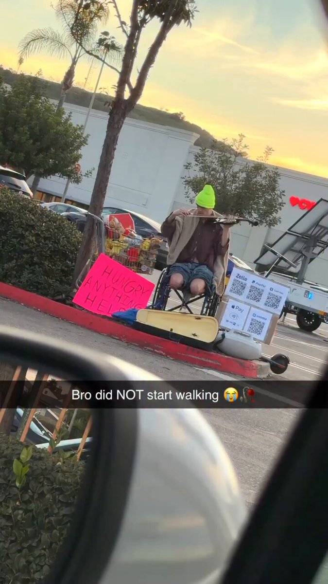 The image shows a person sitting in a wheelchair, holding up signs that say "start walking". The person appears to be making a statement about the importance of walking for one's health or well-being. The scene takes place near a parking lot with cars parked nearby and trees visible in the background.