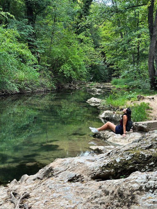 A woman sits peacefully on rocky terrain beside a calm, clear stream surrounded by lush green forest foliage.