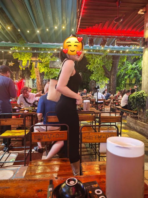 A woman in a black dress walks through an outdoor restaurant at night with string lights and other diners visible in the background, her face covered by a heart-eyes emoji.