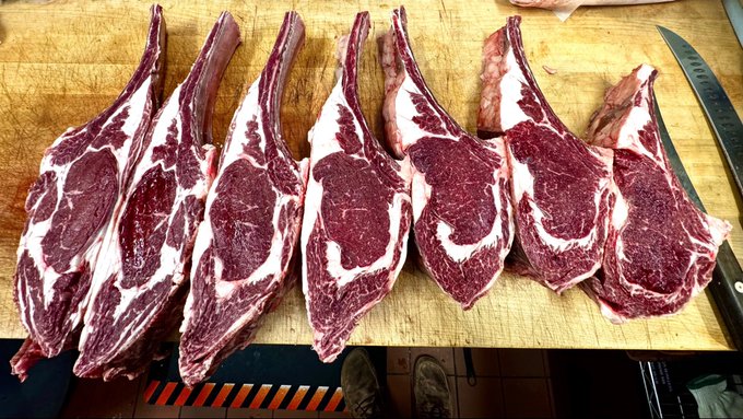 Seven raw tomahawk steaks with prominent bones are arranged in a row on a wooden cutting board, with a knife visible on the right side.