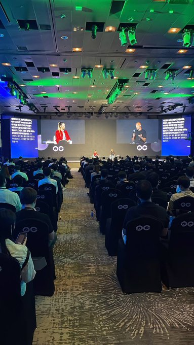 A view from the back of a large conference hall where two speakers appear on dual projection screens, with attendees seated at tables marked with infinity logos.