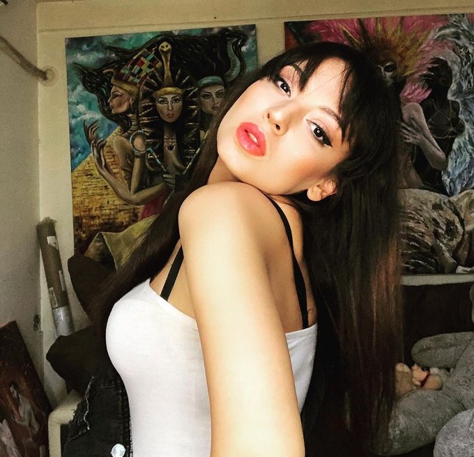 A young woman with long dark hair and a nose ring poses in a white tank top, standing in front of a wall decorated with Egyptian-themed paintings and statues.