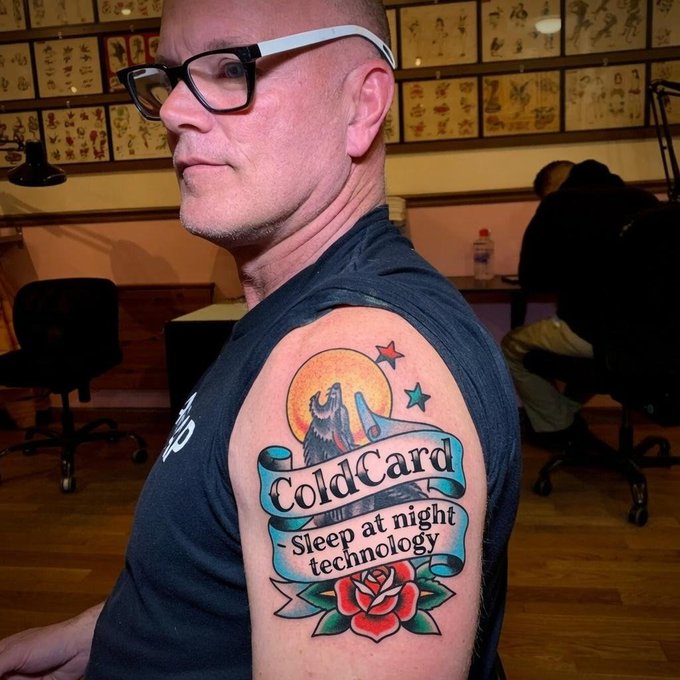 A side profile of a man with glasses showing off a tattoo on his shoulder that reads "ColdCard - Sleep at night technology" featuring a wolf and rose design.