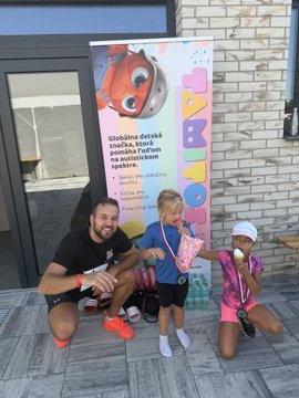 A man and two young girls pose together on a paved sidewalk next to a building, with one girl wearing a pink outfit and white cap, the other in a blue shirt, and a colorful banner behind them that reads 'ZABAVKO' and 'Stalčno detski značka, która pomože tvojim na zúčastniť sa života v...'.