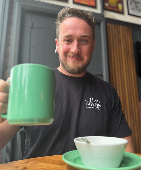 A smiling bearded man wearing a black "Pop's Tavern" t-shirt holds up a large green mug while a bowl of soup sits on the wooden table in front of him.