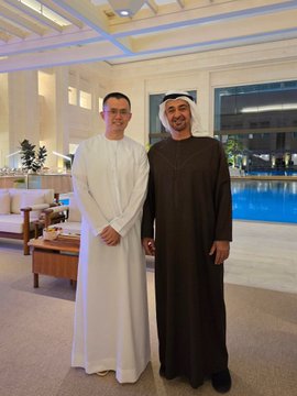 The image features two men dressed in traditional Arab clothing, standing next to each other with their hands together. They are posing for a photo in front of a couch and a coffee table. The background includes a window that offers a view of an indoor swimming pool area. The image also contains text elements such as "CZ @ cz_binance has spent years doing the part of crypto building most people don't see Abu Dhabi Asia and many places around the world Meeting leaders regulators and builders Explaining industry opening doors and helping crypto markets be understood".