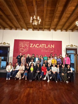 A large group of people poses for a group photo on wooden stairs in front of a red banner that reads "ZACATLÁN, Pueblo Mágico."