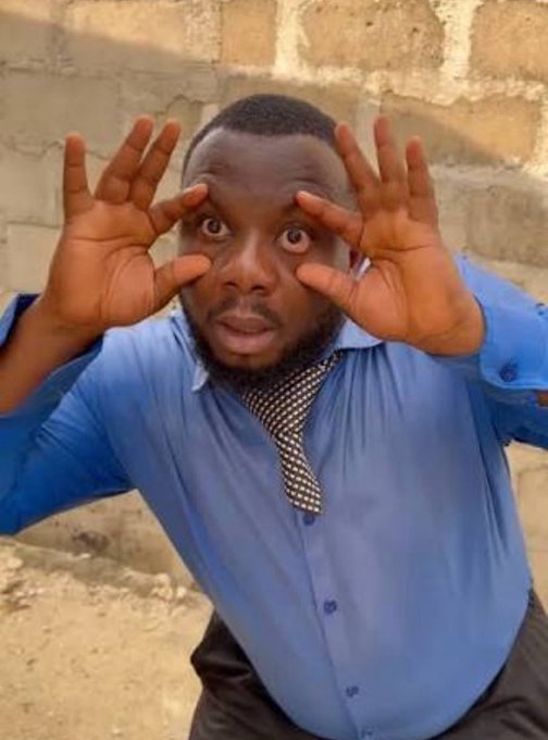 A man in a blue shirt and tie making a surprised expression with his hands framing his face, photographed against a brick wall background.