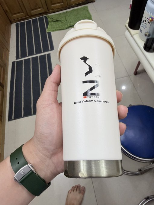 A hand wearing a green-strapped watch holds a cream-colored Zoma Vietnam Community branded tumbler/cup in what appears to be an entryway with floor mats visible in the background.