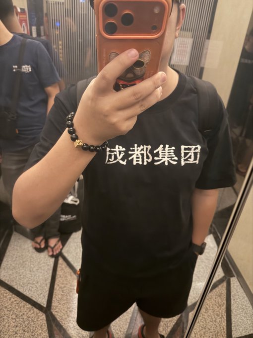 A mirror selfie of a person wearing a black t-shirt with Chinese characters '成都集团' (Chengdu Group) and a bear design, holding a phone with a red case that has a cartoon bear face, also wearing a black beaded bracelet with a gold charm.