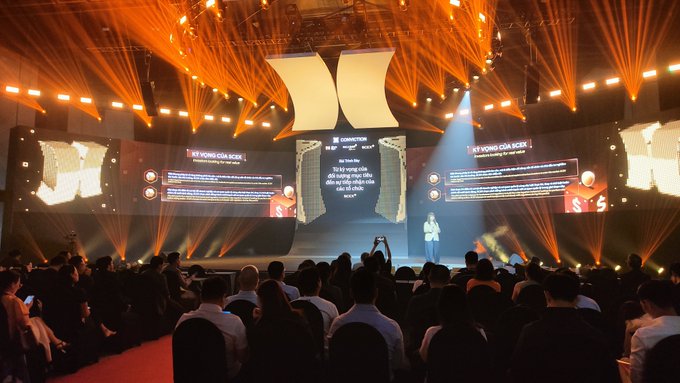 A large conference event with dramatic orange stage lighting where a speaker presents to a seated audience in front of multiple large screens displaying 'KỶ NGUYÊN CỦA KICK' in Vietnamese.