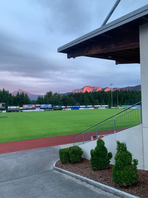 A soccer field nestled among mountains glows at sunset, with advertising banners for "BWT" lining the pitch beneath a modern stadium roof. The serene alpine setting matches the upbeat energy of a "good morning" greeting heralding "$RIVER season 6."