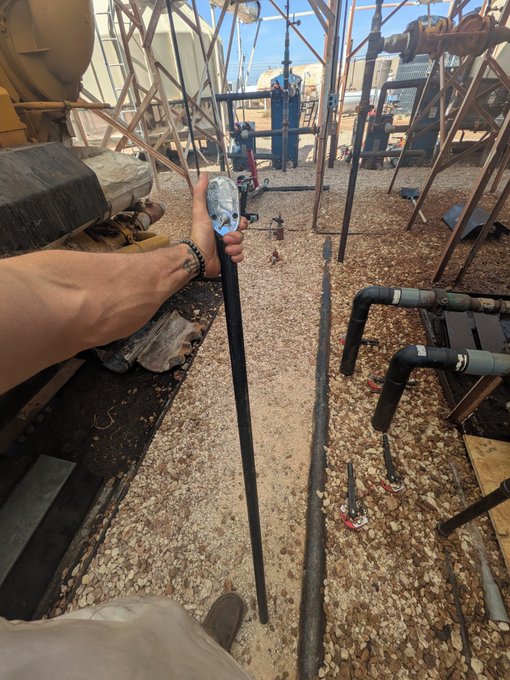 A first-person perspective shows a tattooed arm holding a long rod in an industrial yard filled with gravel, piping, tanks, and metal framework.
