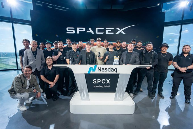 The image features a group of men posing together, with some standing behind a desk or table that has a sign reading "SPACEX" on it. The people are wearing black clothing, which complements the dark background of the photo. In addition to the main group, there is also an individual holding a small object in their hand, possibly a toy or a decoration related to the event.