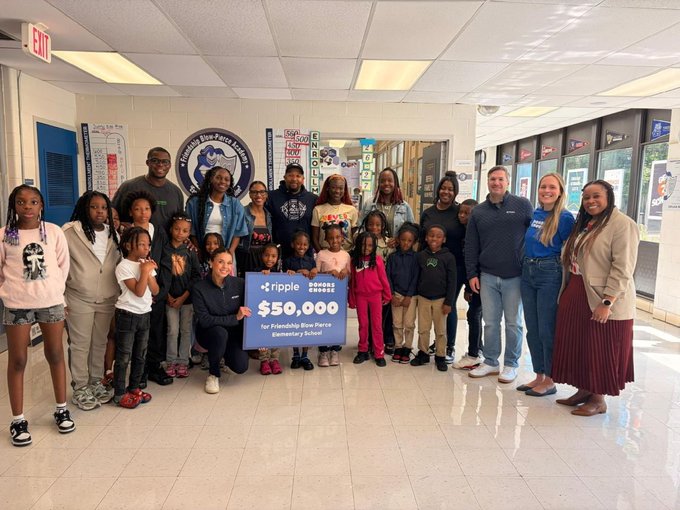 A group of children are posing with their teachers and principal at a school, holding up a blue sign that reads "$50,000 for supplies". The classroom appears to be well-equipped, including desks and chairs arranged neatly in rows, as well as various books scattered around the room. The school is located within a large building with many columns on each floor, suggesting it may have multiple classrooms or offices. The group seems to be gathered for an event, possibly celebrating a successful fundraising campaign that will improve the classroom conditions for the students.