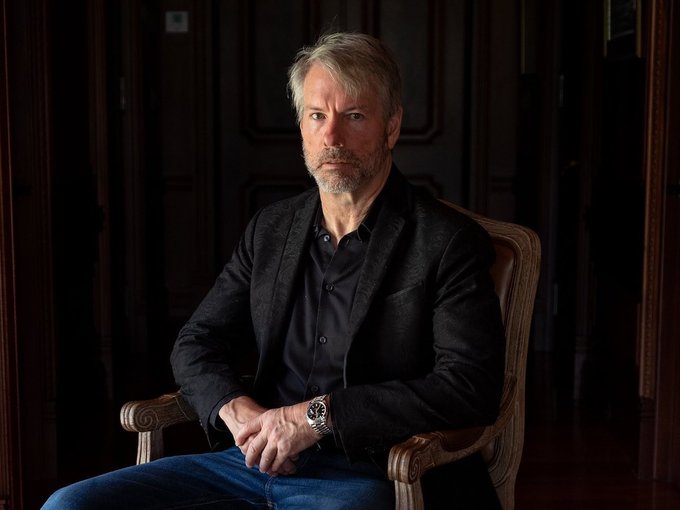 A middle-aged man with graying hair and a beard sits in a wooden chair against a dark paneled background, wearing a black blazer over a dark shirt and a silver wristwatch. The image accompanies a post about Michael Saylor's personal commitment to holding Bitcoin, with his serious demeanor visually reinforcing his statement of unwavering conviction in the cryptocurrency.