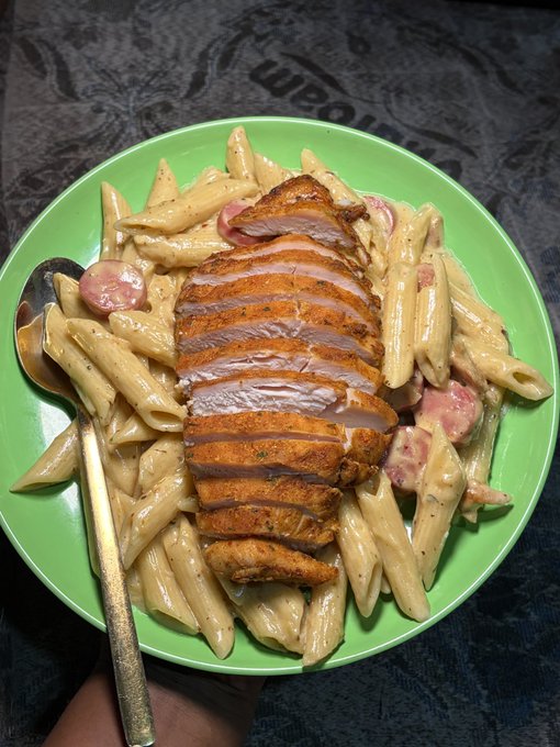 A green plate filled with penne pasta in a creamy sauce mixed with sliced sausage, topped with sliced seasoned chicken breast, with a spoon resting on the side.