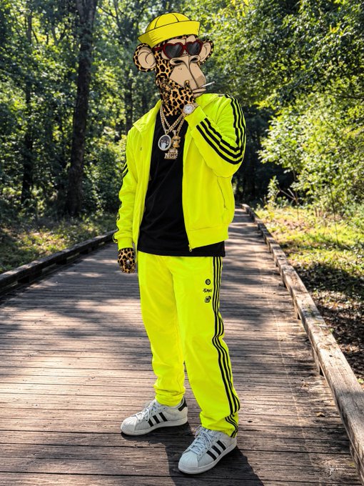 A figure with a leopard-print face, sunglasses, yellow cap, and neon green Adidas tracksuit stands on a wooden boardwalk in a forested area, wearing white sneakers.