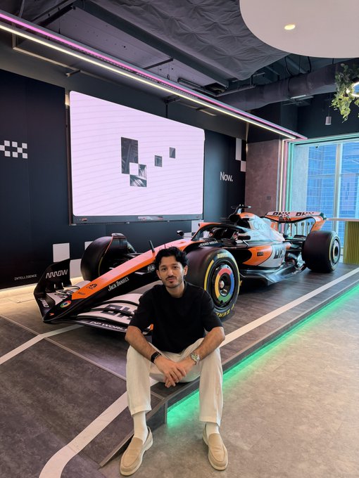 A man in a black shirt and beige pants sits in front of a Formula-style race car (orange and black) inside a showroom with a large display screen and the word "Now."