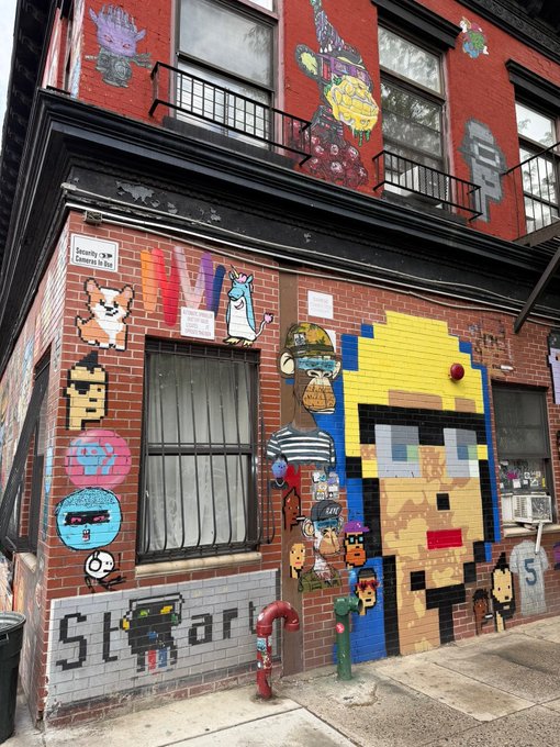 A red brick building corner covered in colorful street art and pixel-art murals, including a yellow cartoon face and various other characters, with a red fire hydrant on the sidewalk.