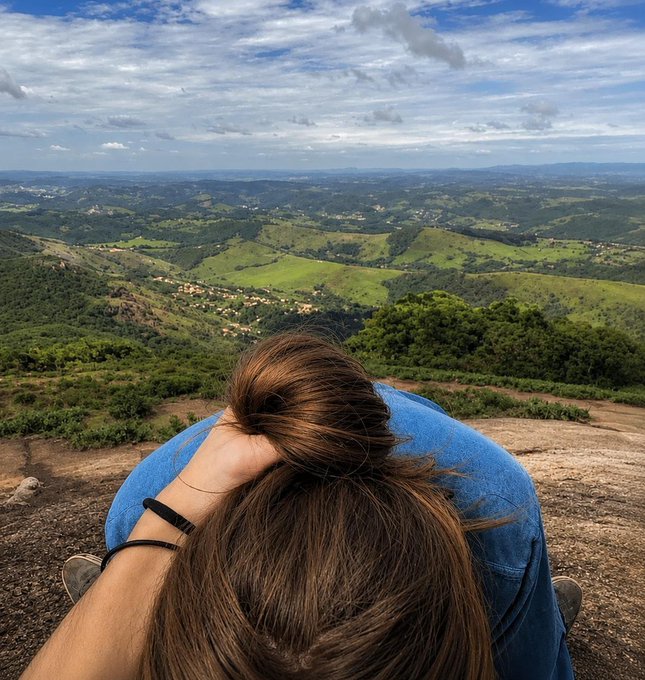 An overhead view of a person with brown hair tied up sitting on a cliff edge, looking out over a vast, green hilly landscape under a partly cloudy sky.