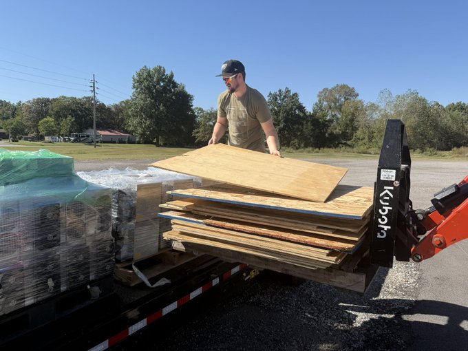 A man wearing a cap and sunglasses unloads wooden boards from a flatbed trailer using a Kubota tractor with a loader attachment.