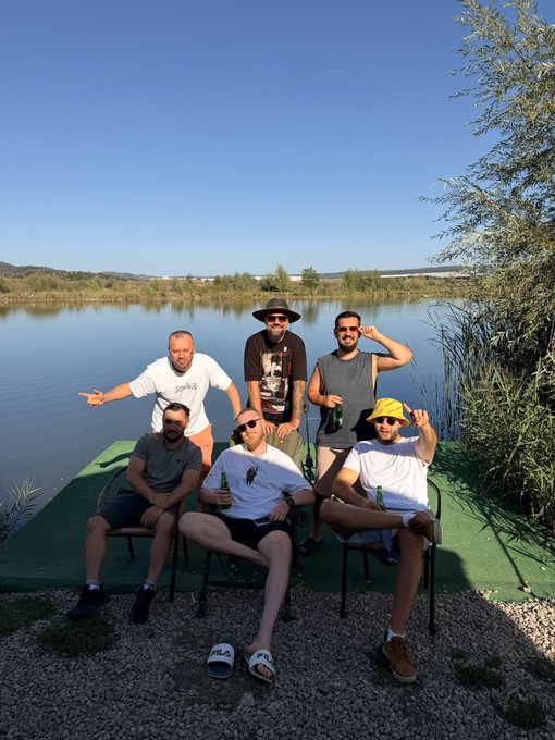A group of six people posing for a photo outdoors on a green platform next to a lake on a sunny day.