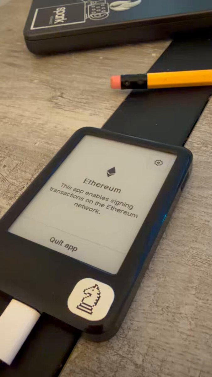 The image shows a black e-reader with a white sticker, placed on top of a wooden table. The e-reader displays an error message that reads "Ethereum transaction failed", indicating that there was an issue with the transaction. A yellow pencil is also present in the scene, lying next to the e-reader.