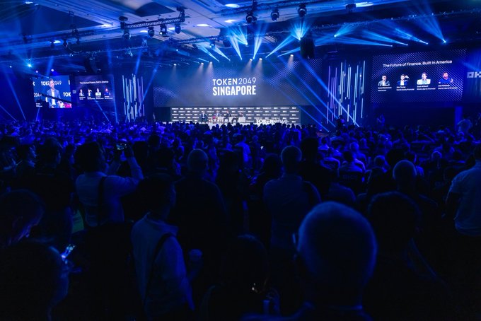 A crowded conference hall lit with blue lights, showing attendees gathered for 'TOKEN2049 SINGAPORE' with the tagline 'The Future of Finance, Built in AI America' on the screens.