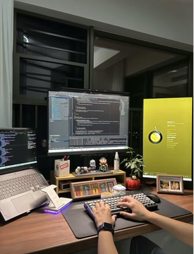 A multi-monitor coding workspace at night with a person typing on a mechanical keyboard, featuring figurines and a clock on the desk.