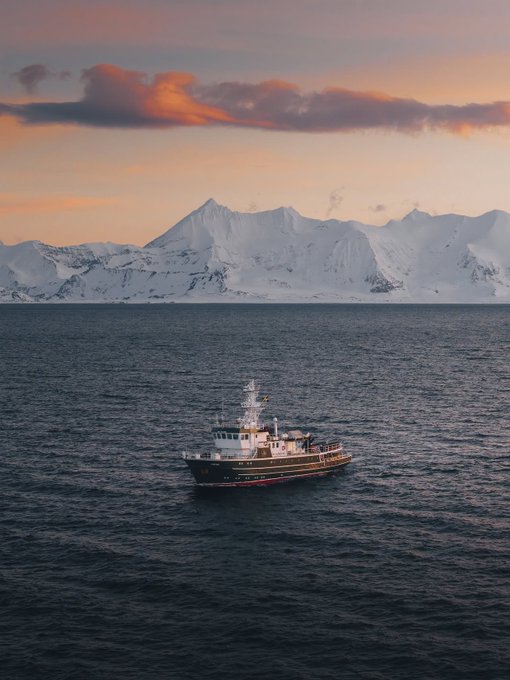 A small boat floats on dark ocean waters with a dramatic, snow-covered mountain range in the background under a cloudy sky.