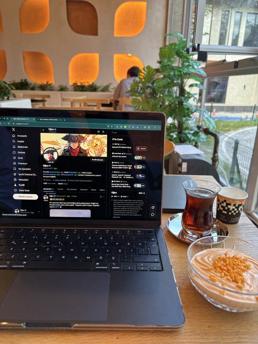 In this image, a laptop with various social media applications open is placed on a dining table next to a glass bowl of cereal topped with nuts, a spoon beside the bowl, and two cups - one containing tea and another filled with coffee. A person can be seen in the background near a plant. The scene takes place at a café where the person appears to be working or browsing social media while enjoying their breakfast.