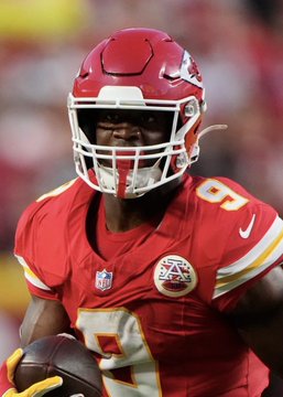 The image shows a Kansas City Chiefs player wearing a red jersey with the number 9 and a matching helmet, holding a football while wearing yellow gloves; this relates to the accompanying post about betting on Kenneth Walker to score a touchdown, as Walker is a running back, though the player pictured appears to be from the Chiefs rather than Walker's Seahawks team.