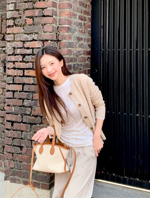 A woman with long dark hair stands smiling against a brick wall, wearing a beige cardigan, white top, and cream skirt while holding a structured handbag.