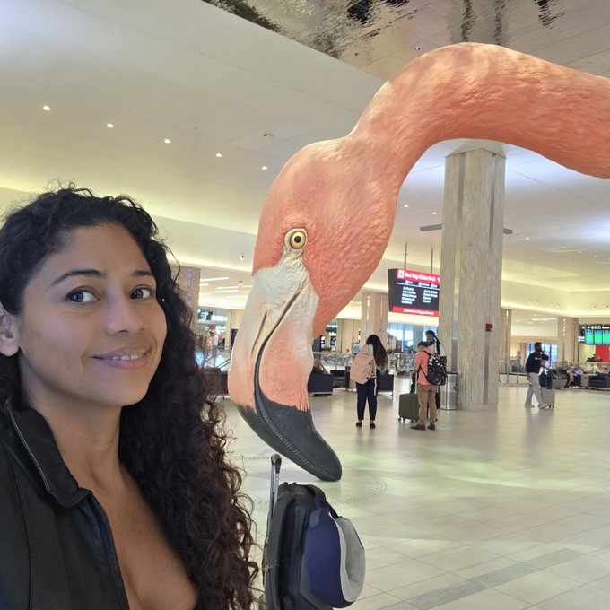 A smiling woman takes a selfie in an airport terminal next to a large pink flamingo statue.